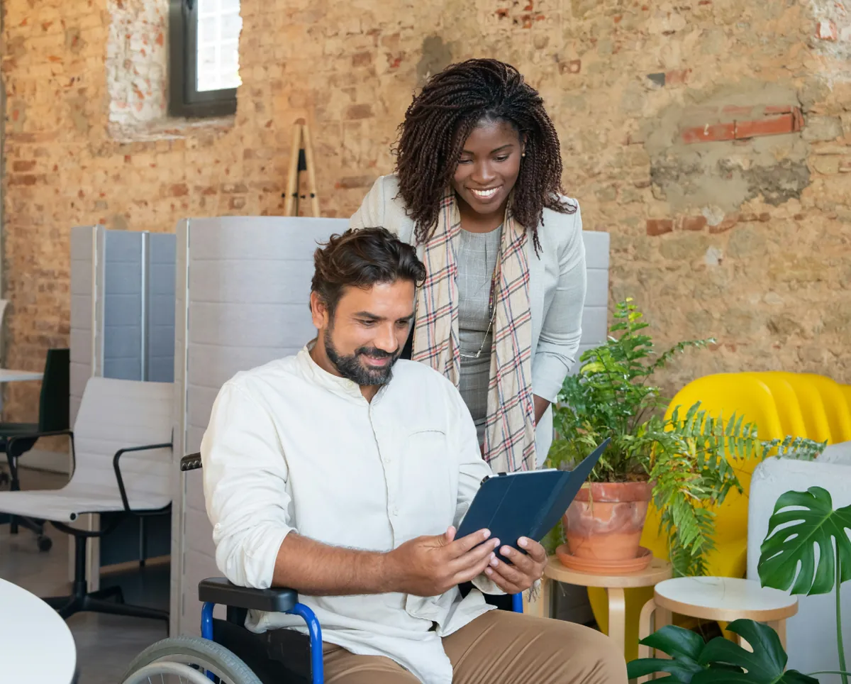 A support worker smiles as a man in a wheelchair reviews information on a tablet in a bright office